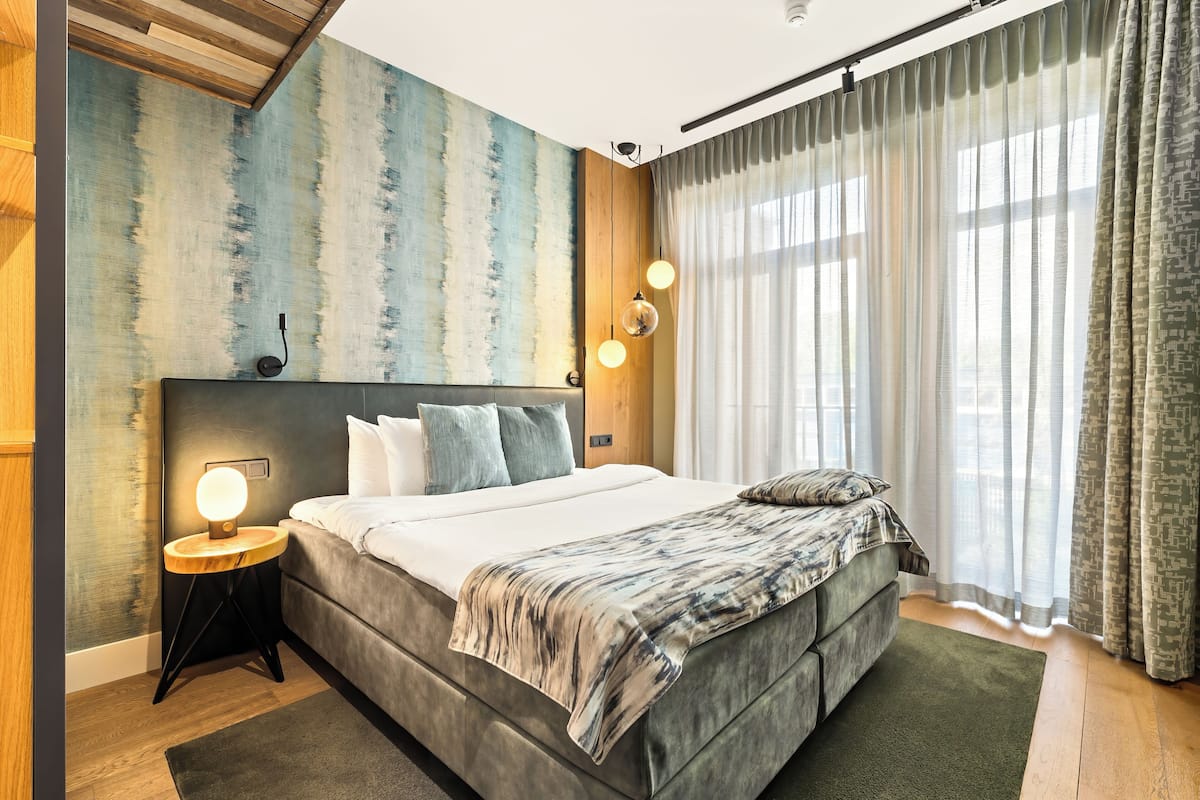 A modern bedroom with a large bed, a nightstand with a lamp, a wooden headboard, and a large abstract painting on the wall.