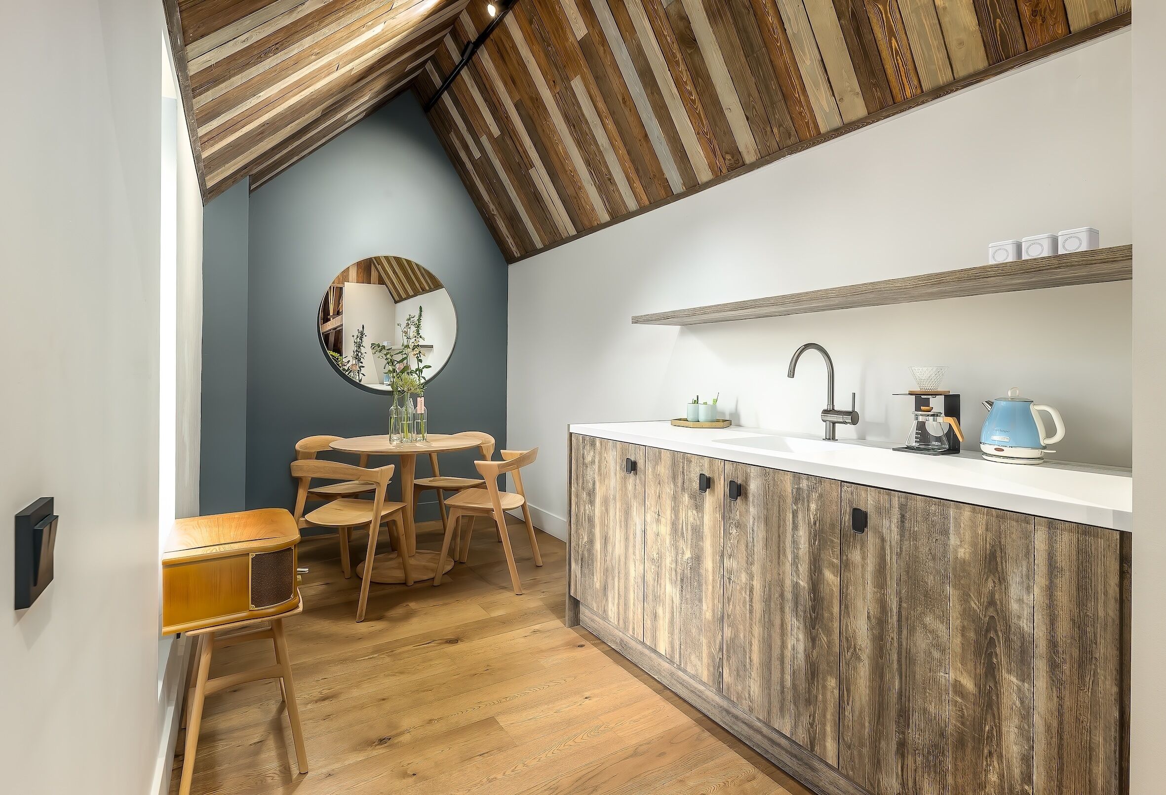 Una cocina con techo de madera, un espejo redondo, una mesa de comedor con sillas y una encimera blanca con un lavamanos y armarios.