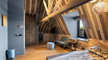 Une chambre à coucher aménagée dans un espace de type « loft », avec des poutres apparentes, un lit, un bureau et une fenêtre.