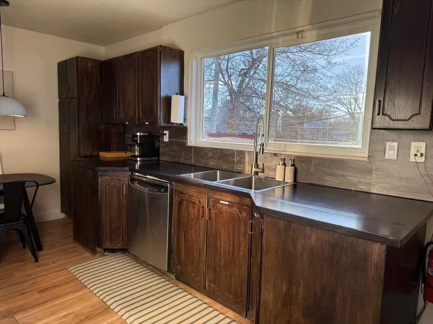 Una cucina moderna con mobili in legno scuro, lavastoviglie in acciaio inossidabile, un lavello a due vasche e una finestra con vista sugli alberi.