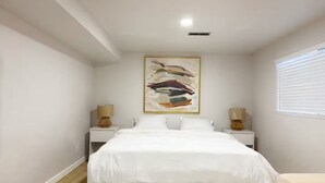 Quarto com cama, duas mesas de cabeceira com luminárias, um quadro abstrato grande e uma janela com persianas.