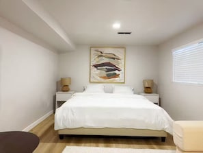 Ein Schlafzimmer mit einem Bett, zwei Nachttischen mit Lampen, einem großen abstrakten Gemälde und einem Fenster mit Jalousien.