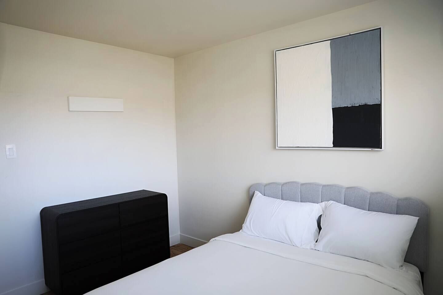 A bedroom with a bed, a black headboard, a white mattress, and a wall-mounted artwork.