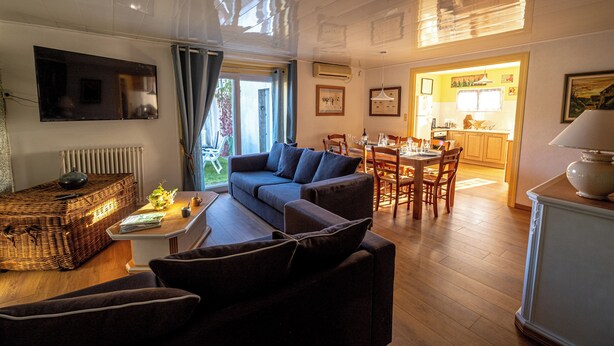 A living room with a sofa, a coffee table, and a television mounted on the wall. There is a dining area with a table and chairs, and a kitchen visible through an open door.