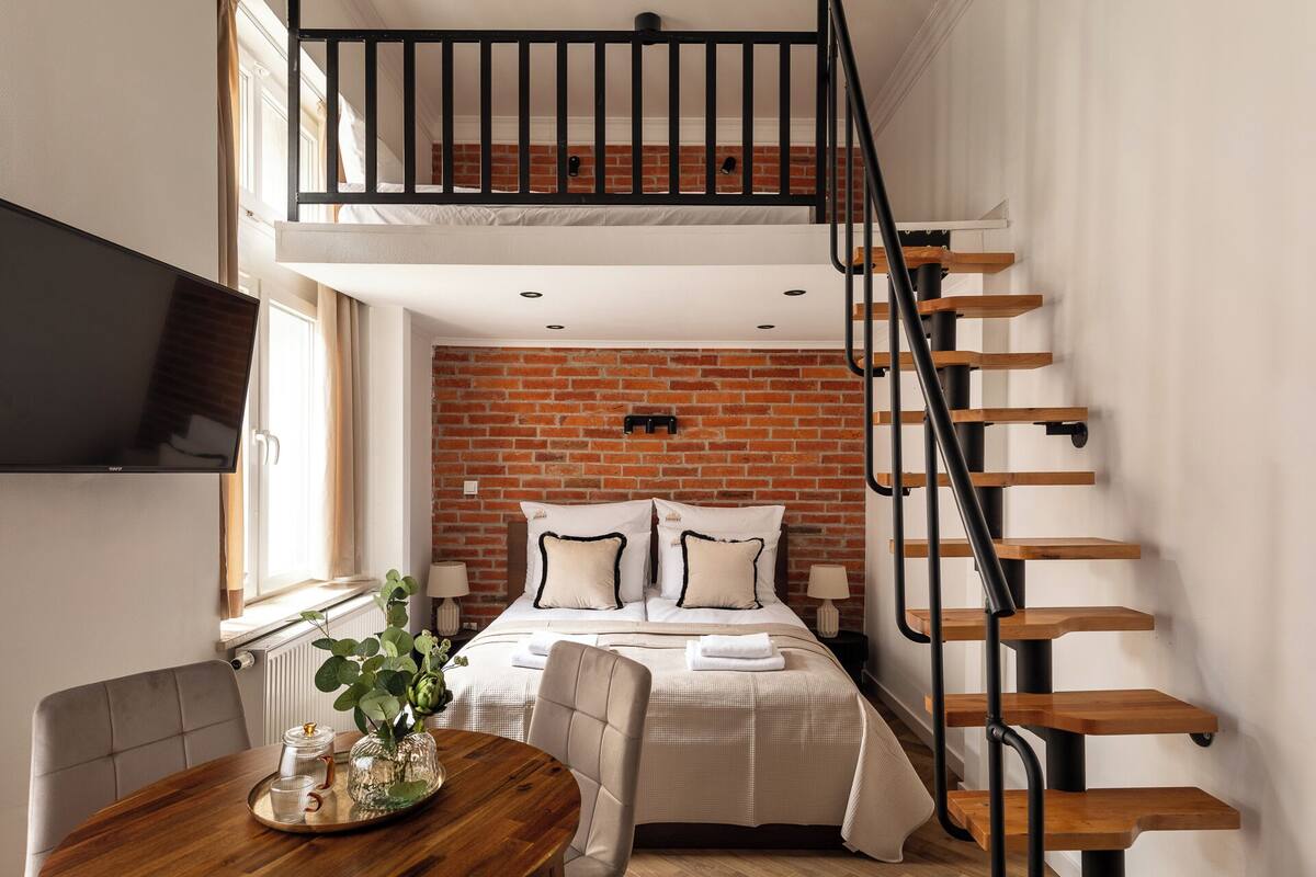 Un dormitorio estilo loft con pared de ladrillo, cama con ropa de cama blanca, mesa de comedor de madera con un jarrón y escalera con peldaños de madera y barandales negros.