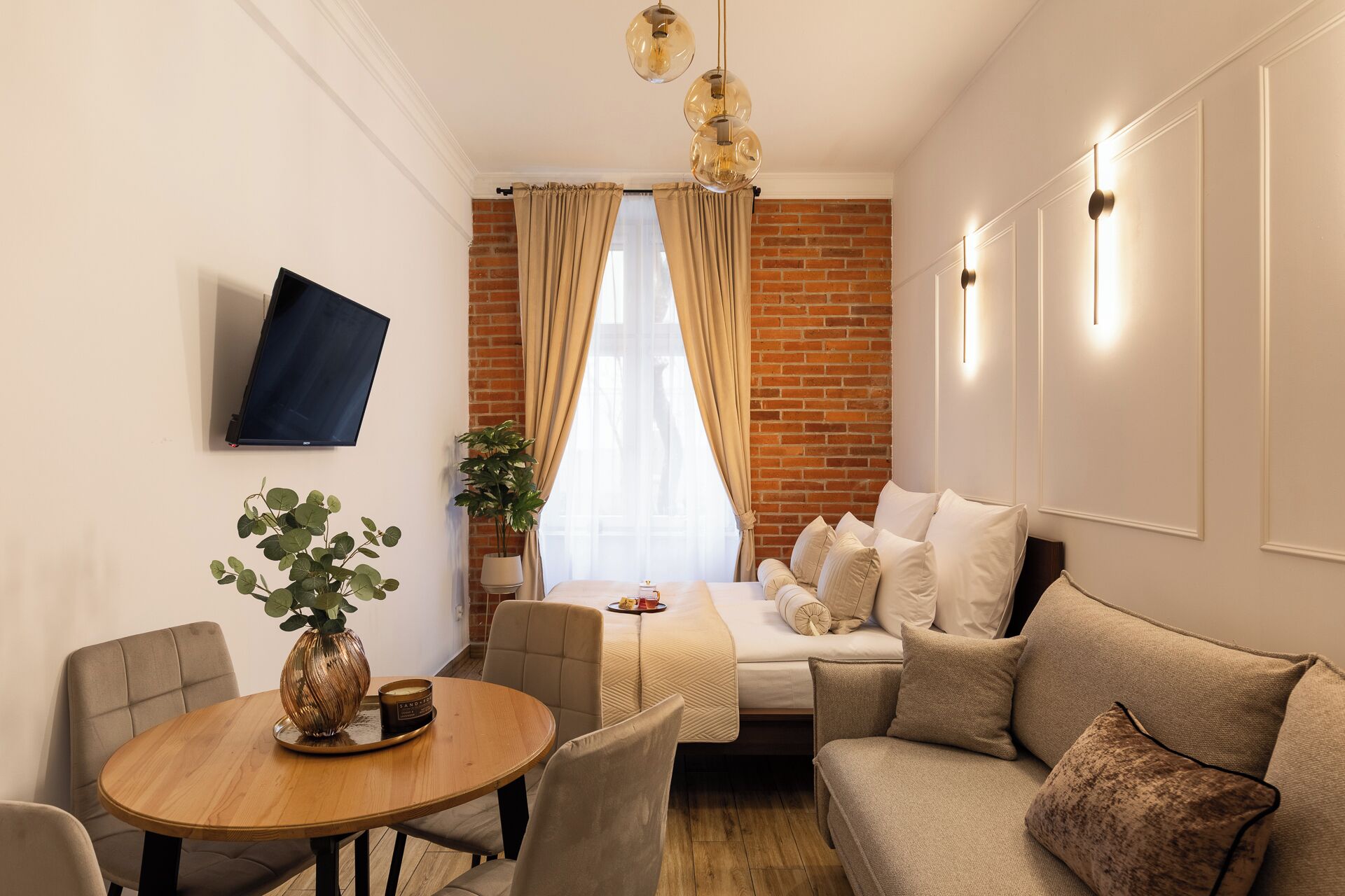 A modern living room with a sofa, a dining table with chairs, a TV mounted on the wall, and a brick wall.