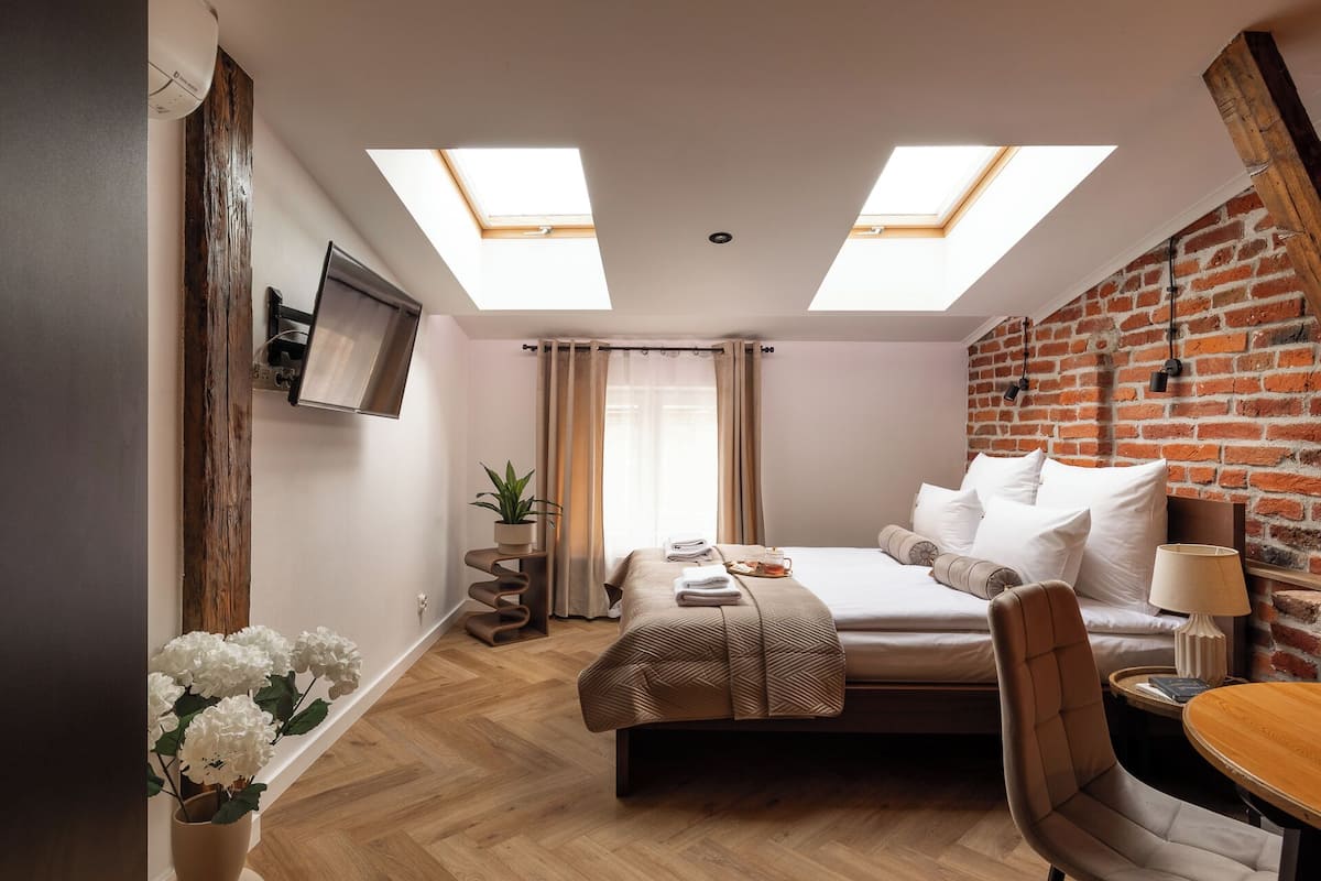 Una habitación de hotel moderna con una cama, un escritorio y una silla. Hay tragaluces en el techo, una pared de ladrillo y un televisor montado en la pared.