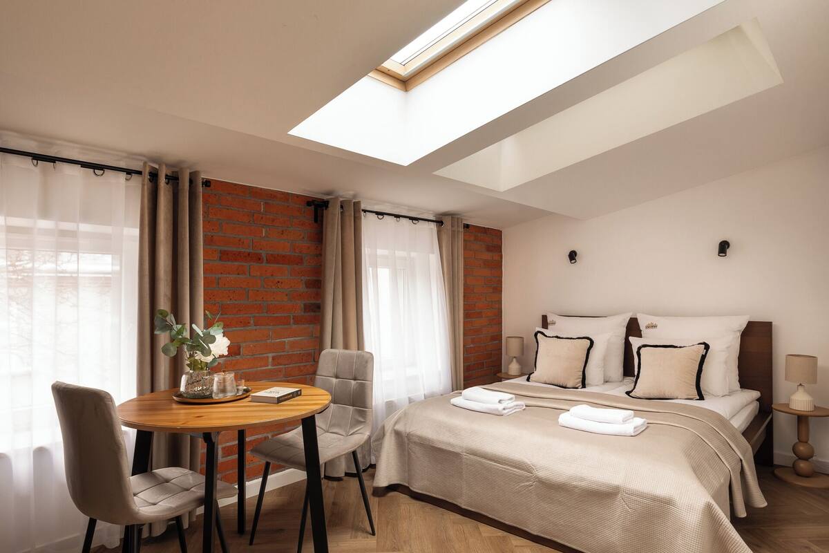 Una habitación de hotel con tragaluz, una cama con ropa de cama blanca, una mesa de madera con un florero y una silla.