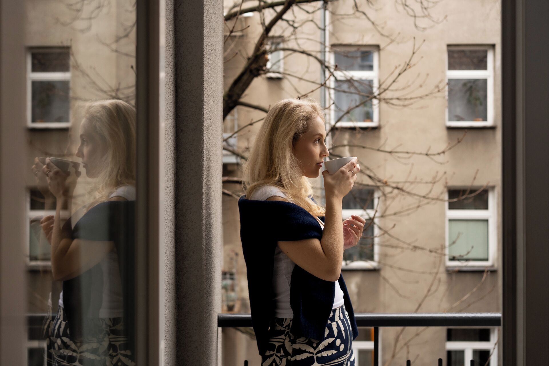 A person standing by a window with a view of a building with multiple windows and bare trees.
