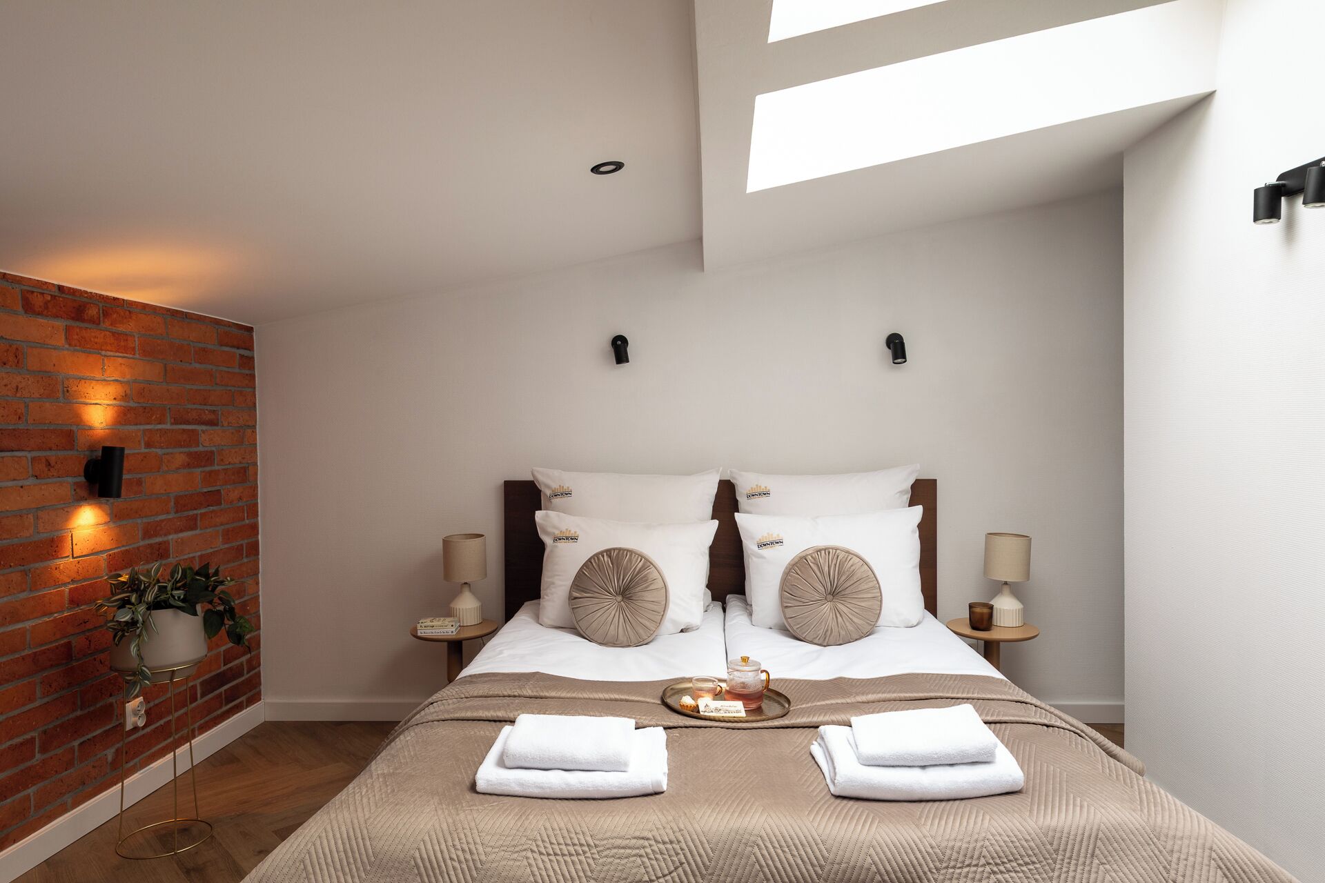 Een moderne hotelkamer met een accentmuur van baksteen, een groot bed met een beige sprei, twee nachtkastjes met lampen en een kleine potplant.