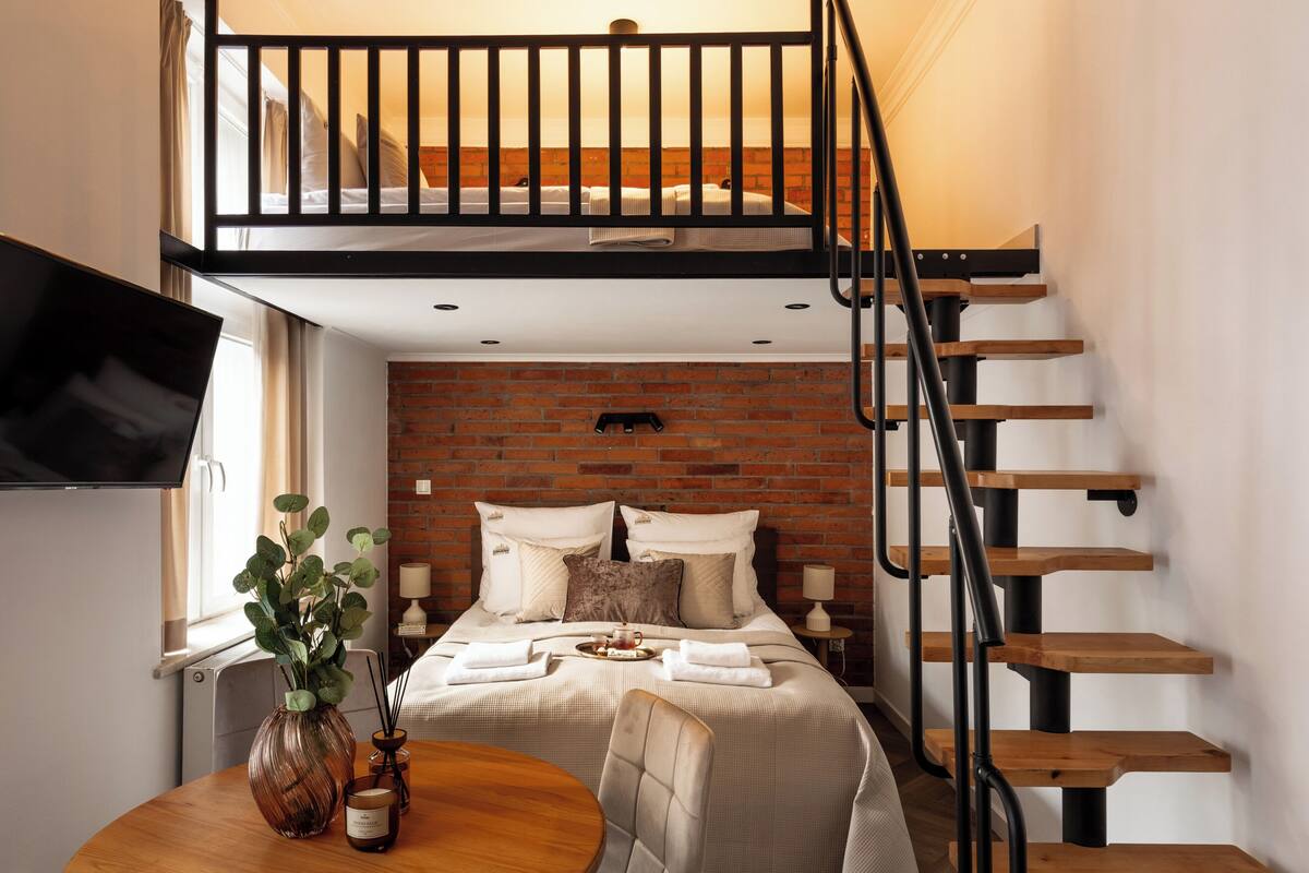 Un dormitorio con cama de altillo, pared de ladrillo, escalera de madera, televisor, mesa de comedor y un florero con plantas.