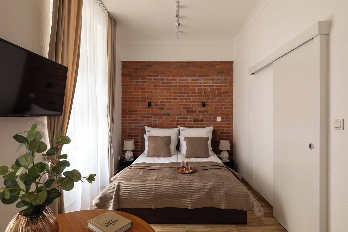 Habitación de hotel con pared de ladrillo, una cama con colcha de tonos beige y marrón, dos lámparas de noche, un televisor de pantalla plana montado en la pared y una mesita con un libro y una bandeja con tetera y vasos.