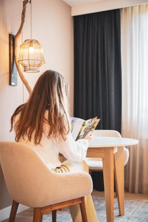 Una persona sentada a una mesa leyendo una revista en una habitación con una lámpara, cortinas y una silla.