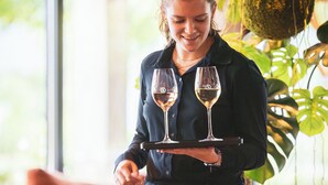 A person holding a tray with two wine glasses, standing in a room with a large plant and a brown sofa. - Hotel de Sterrenberg (Otterlo)