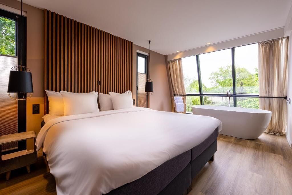 A modern bedroom with a large bed, a bathtub, and a view of greenery through large windows.