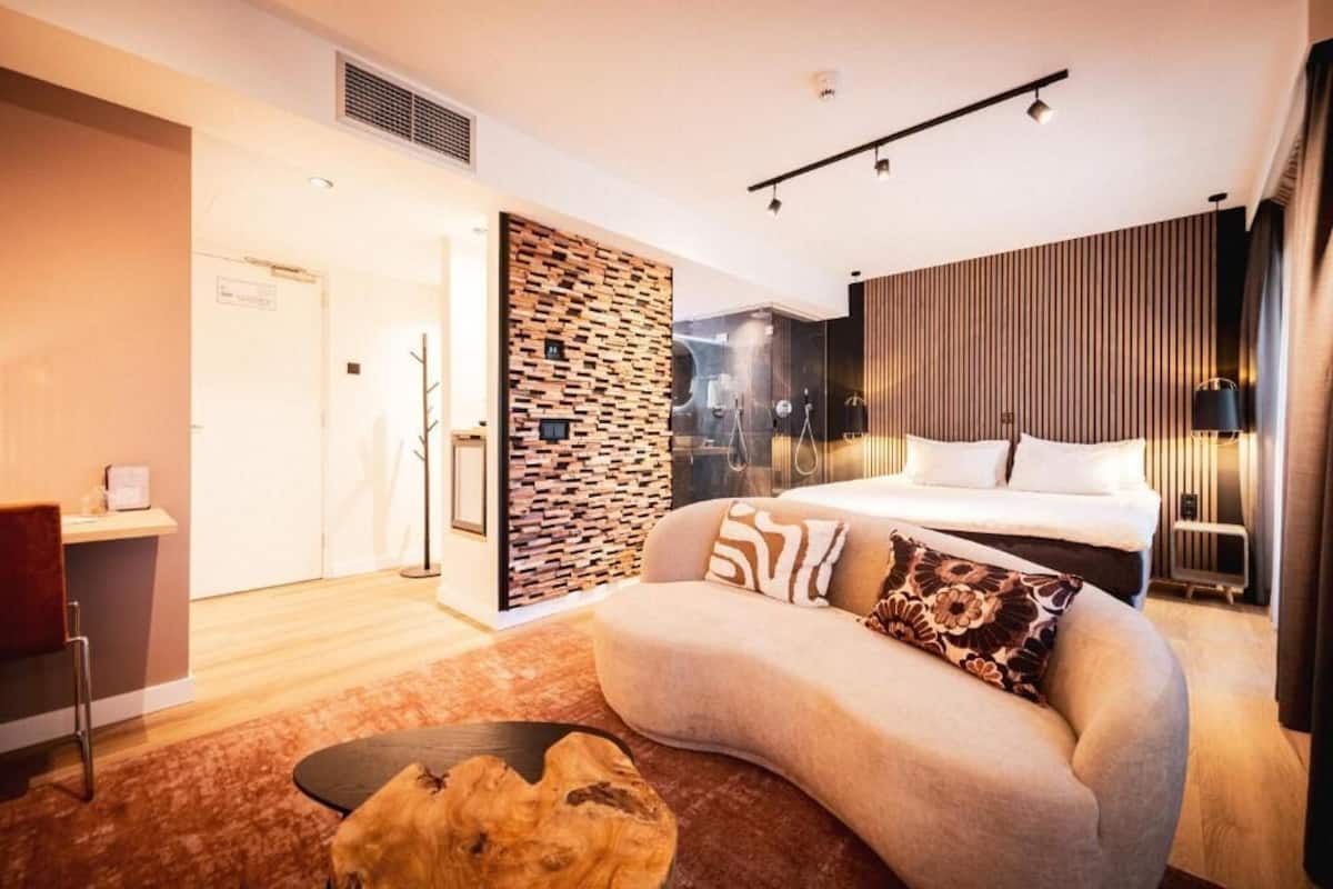 Ein modernes Hotelzimmer mit einer gebogenen Couch, einem Bett mit weißen Leinen, einer Wand aus Holzpaneelen und einem großen Fenster.