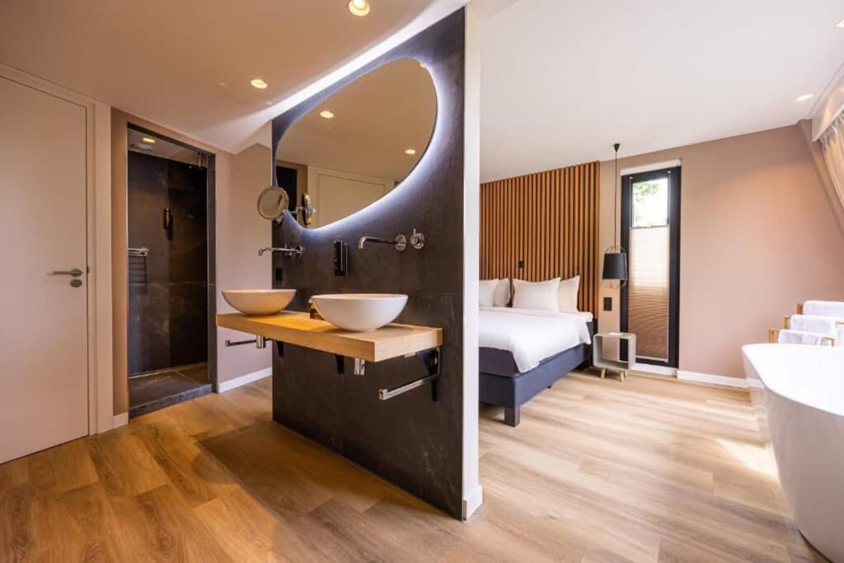 Ein modernes Hotelzimmer mit einem großen Bett, einem Waschtisch mit zwei Becken, einem großen Spiegel und einem separaten Badezimmerbereich.