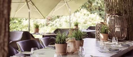 An outdoor dining area with a long table set for two, surrounded by chairs, potted plants, and a large umbrella providing shade.