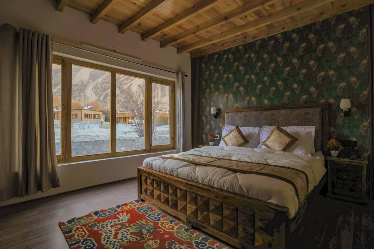 A bedroom with a large window, wooden beams, patterned wallpaper, and a bed with multiple pillows.