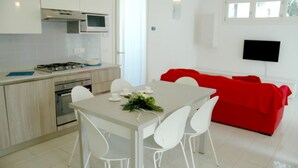 Een moderne keuken met witte kastjes, een gasfornuis en een magnetron. Er is een eettafel met witte stoelen en een rode bank in de woonkamer.