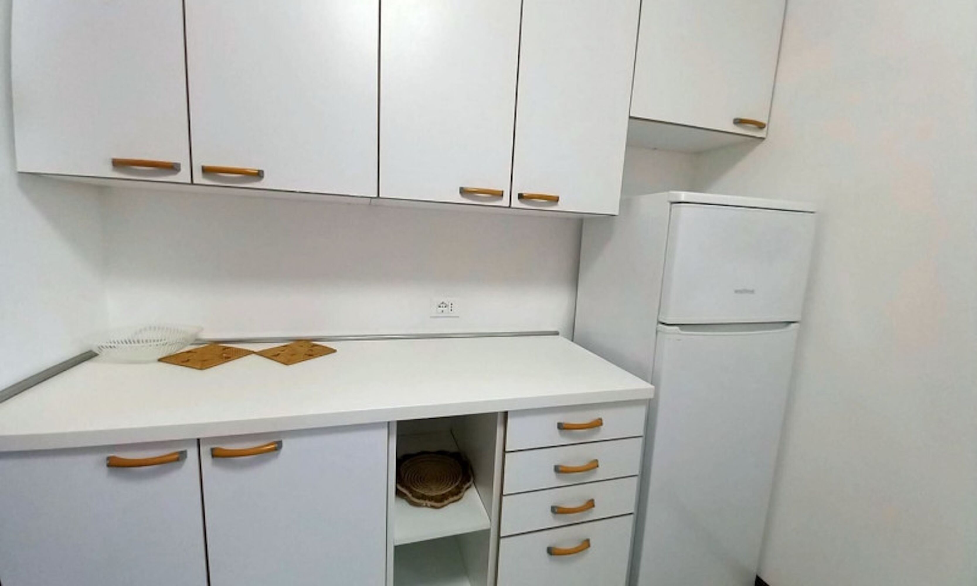 A compact kitchen with white cabinets, wooden handles, and a built-in refrigerator.