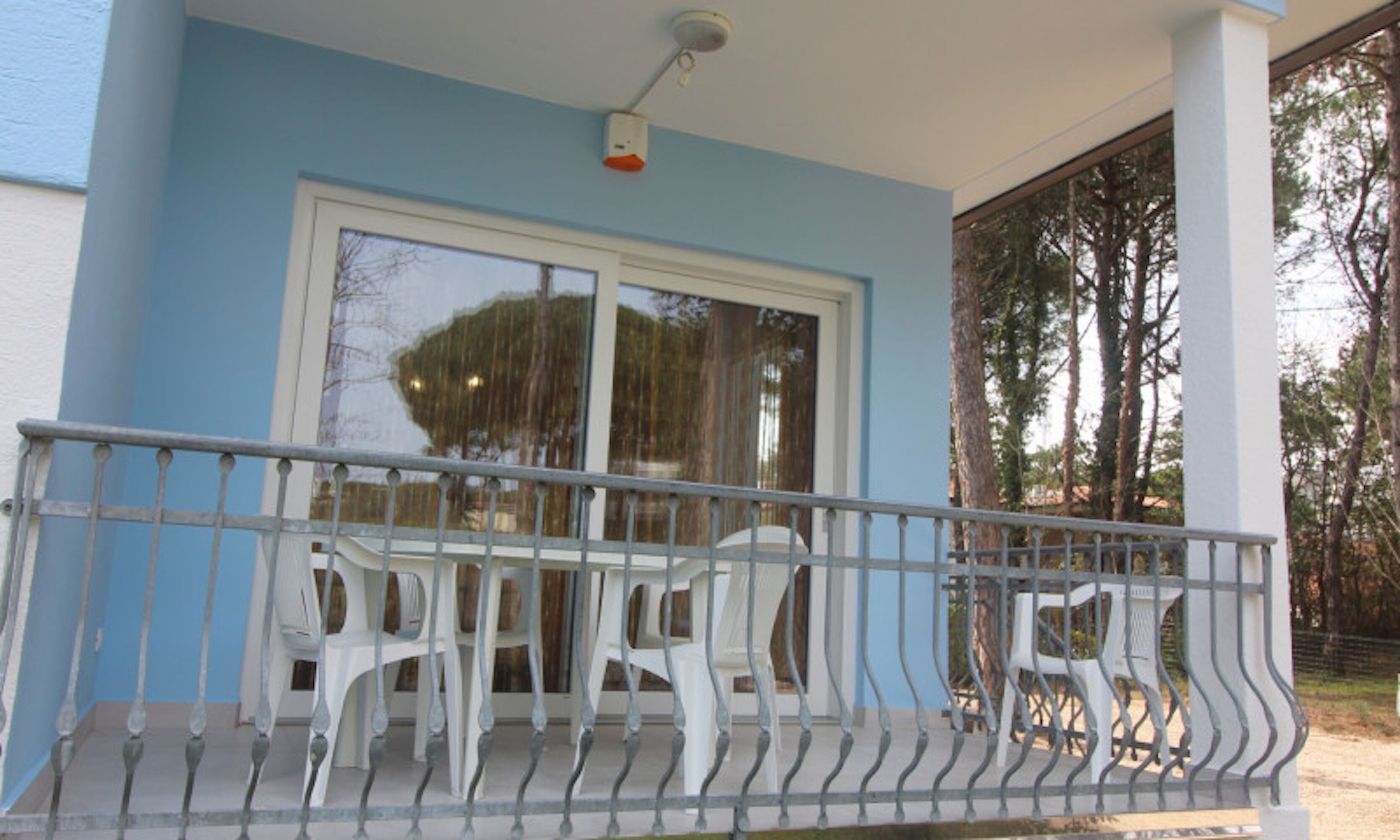 A balcony with a metal railing, two white plastic chairs, and a large window with a view of trees.