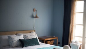 Een slaapkamer met een bed, kussens, een nachtkastje, een lamp, een raam met gordijnen en een hoed op het bed.