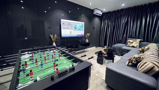 A modern living room with a foosball table, a flat-screen TV, dark curtains, and a sofa with patterned cushions. - The Linkview Shortlet Apartment (Lekki)