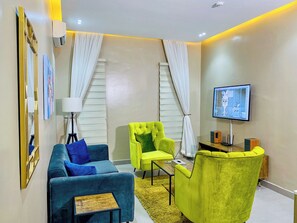 A modern living room with a sofa, armchair, TV, and air conditioning unit. - The Linkview Shortlet Apartment (Lekki)