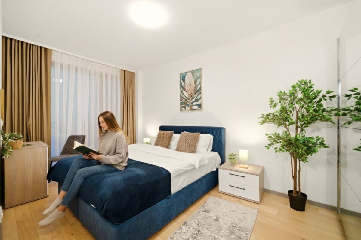 Une chambre à coucher moderne, avec un lit, des tables de chevet, un fauteuil de lecture, une plante en pot et une fenêtre avec des rideaux.