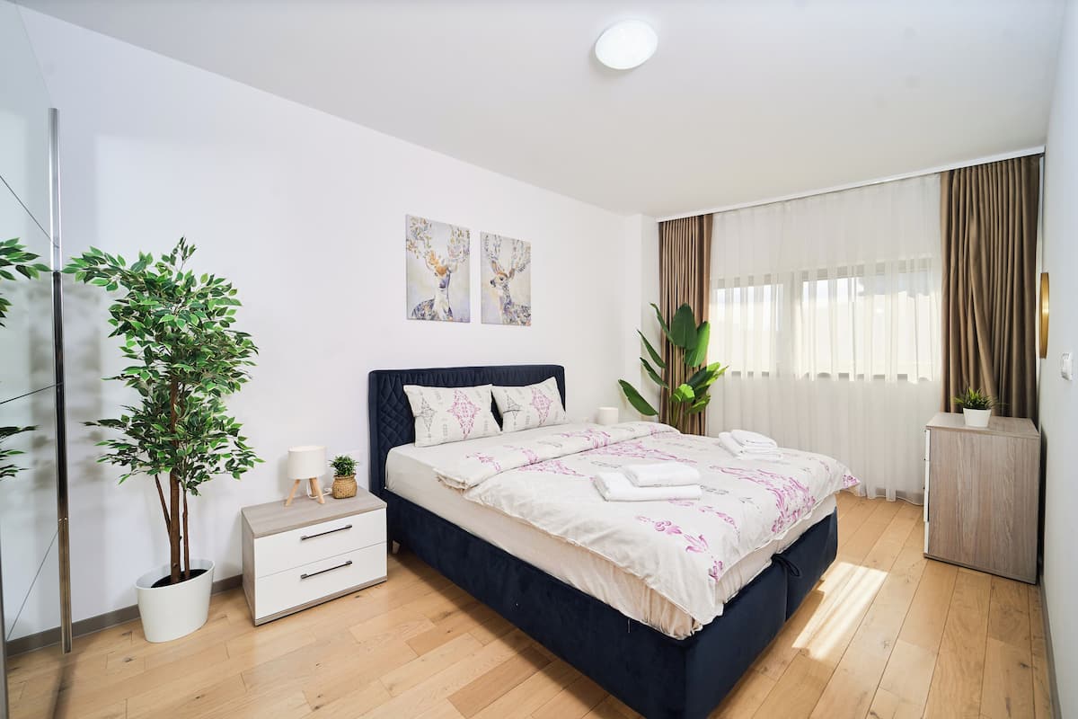 Une chambre à coucher moderne, avec un lit, des tables de chevet, une plante en pot, des œuvres d’art accrochées au mur et une fenêtre avec des rideaux.