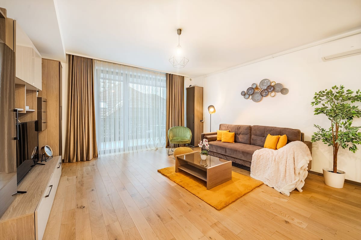 Un salon moderne avec un plancher en bois, un canapé marron agrémenté de coussins jaunes, une table basse en verre, un fauteuil vert et une plante en pot.