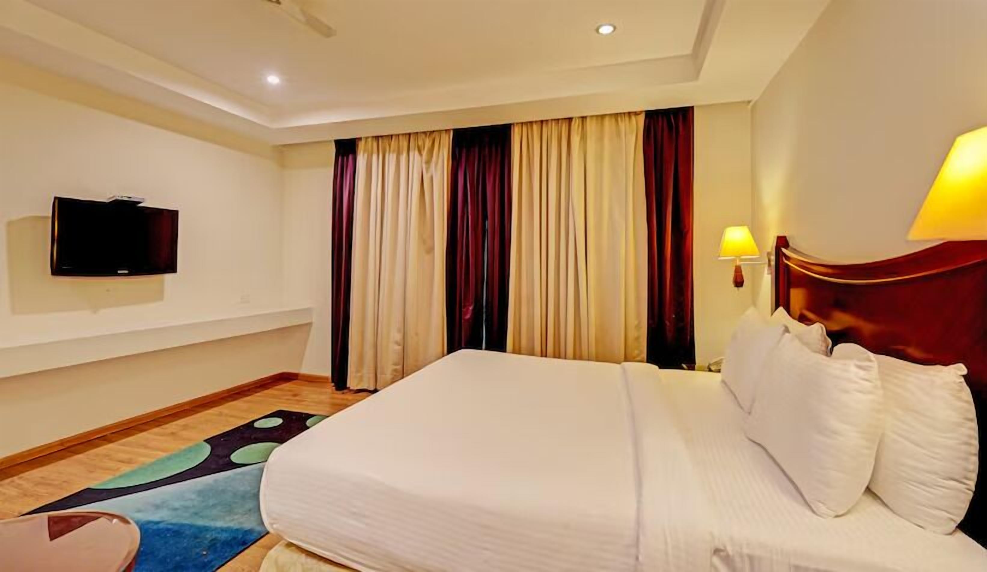 A hotel room with a large bed, a flat-screen TV mounted on the wall, a bedside table with a lamp, and a window with curtains.
