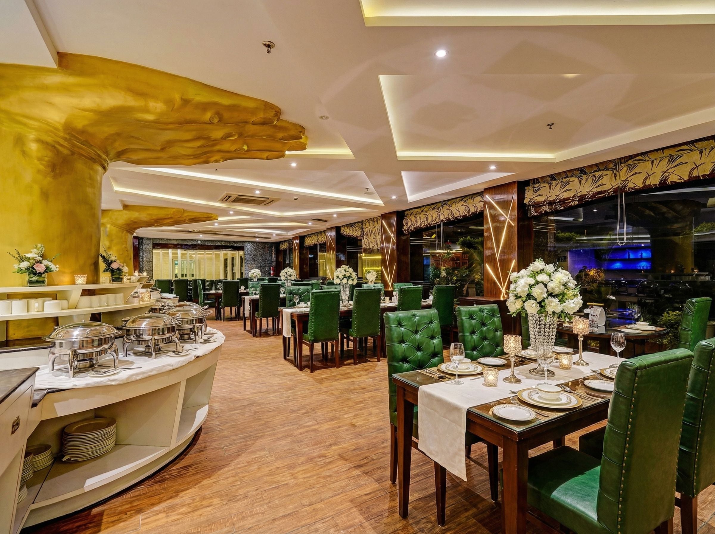 A restaurant with green upholstered chairs, white tablecloths, and a buffet area with silver serving dishes.