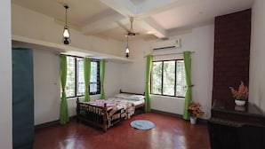 Quarto com cama, duas janelas com cortinas verdes, ventoinha no teto e parede de tijolos.