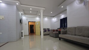 Uma sala de estar moderna com um sofá cinzento grande, uma mesa de centro e uma televisão. Há um pilar branco, um ventilador de teto e uma porta que dá para outro cômodo.