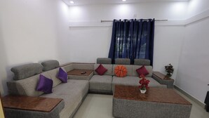Uma sala de estar com um sofá seccional cinza, duas mesas de centro de madeira e uma pequena mesa lateral.