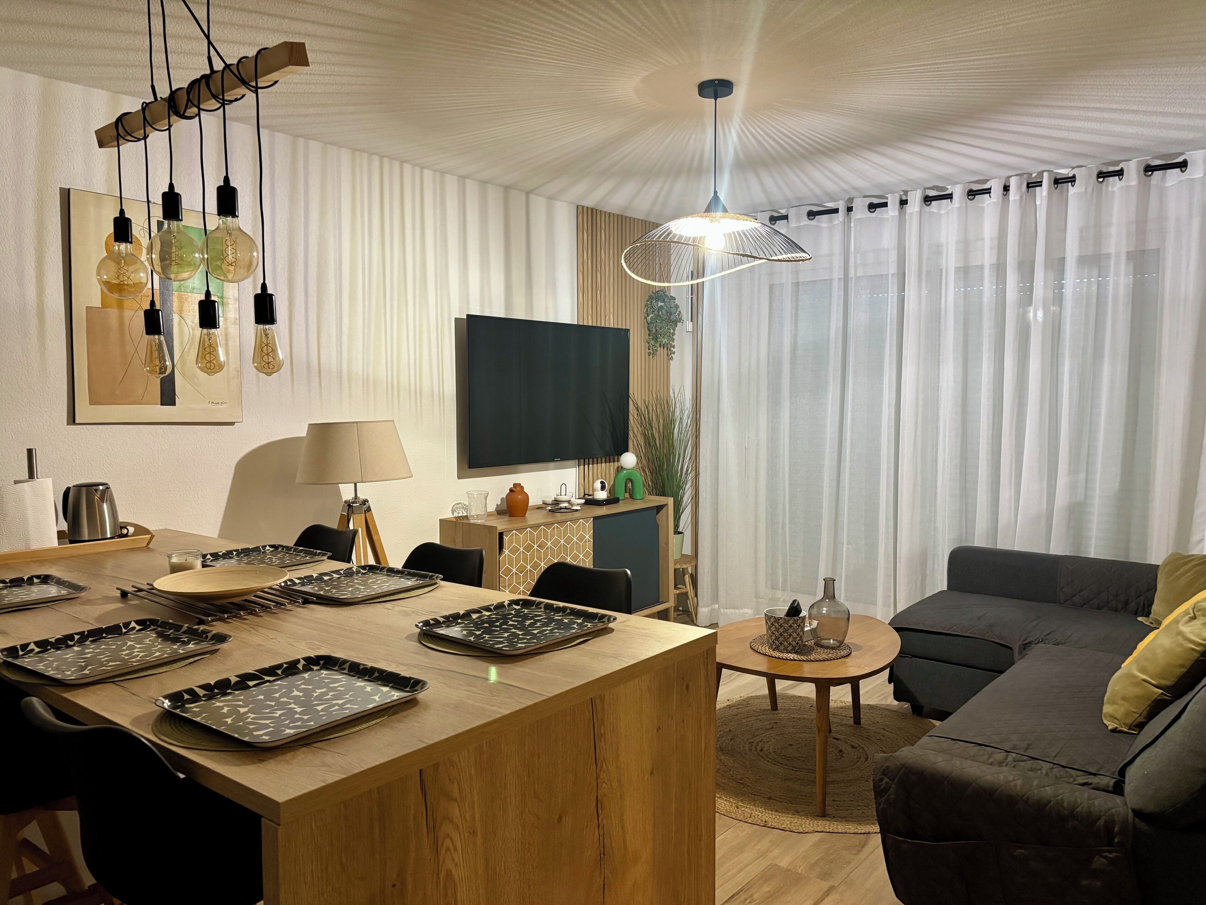 A modern living room with a wooden dining table, black chairs, a grey sofa, and a TV mounted on the wall.