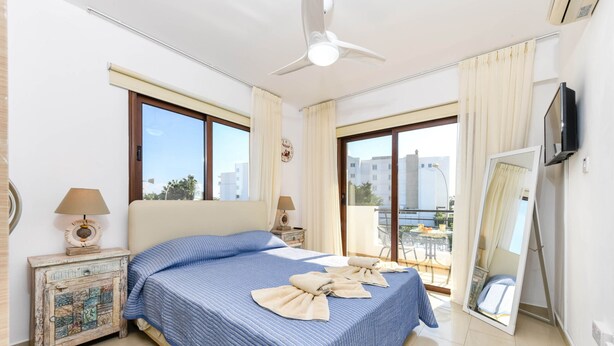 Une chambre d’hôtel avec un grand lit, deux tables de chevet avec des lampes, un ventilateur de plafond, une télévision et un balcon donnant sur des immeubles.