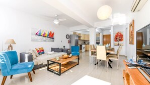 Un salon moderne avec un canapé, un fauteuil bleu et une table basse. On y trouve également un coin repas avec une table et des chaises. Une cuisine est visible au fond.