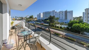 Un balcon avec une table et des chaises, donnant sur une rue bordée de palmiers et sur un immeuble portant l’inscription « Yasmin ».