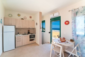 A compact kitchen with a refrigerator, microwave, and sink. There's a dining area with a table and chairs, and a view of the sea through a door. - Mare Blu 2 on the beach (Gennadi)