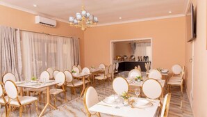 A dining area with multiple tables and chairs, an air conditioning unit, a chandelier, and a television mounted on the wall. - Lavish Suites (Blantyre)