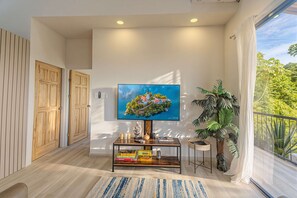Een moderne woonkamer met een flatscreen televisie aan de muur, een houten deur, een kleine tafel met gezelschapsspellen en een grote kamerplant.