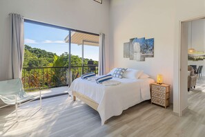 Une chambre avec une grande fenêtre, un lit recouvert de draps blancs, une table de chevet avec une lampe et un tableau accroché au mur.