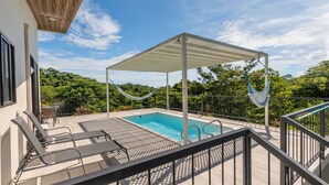 Un área de piscina con una zona de estar techada, una hamaca y vistas a un paisaje boscoso.