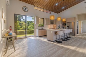 Une cuisine moderne dotée d’un îlot central, de luminaires suspendus et offrant une vue sur la verdure grâce à de grandes portes coulissantes.