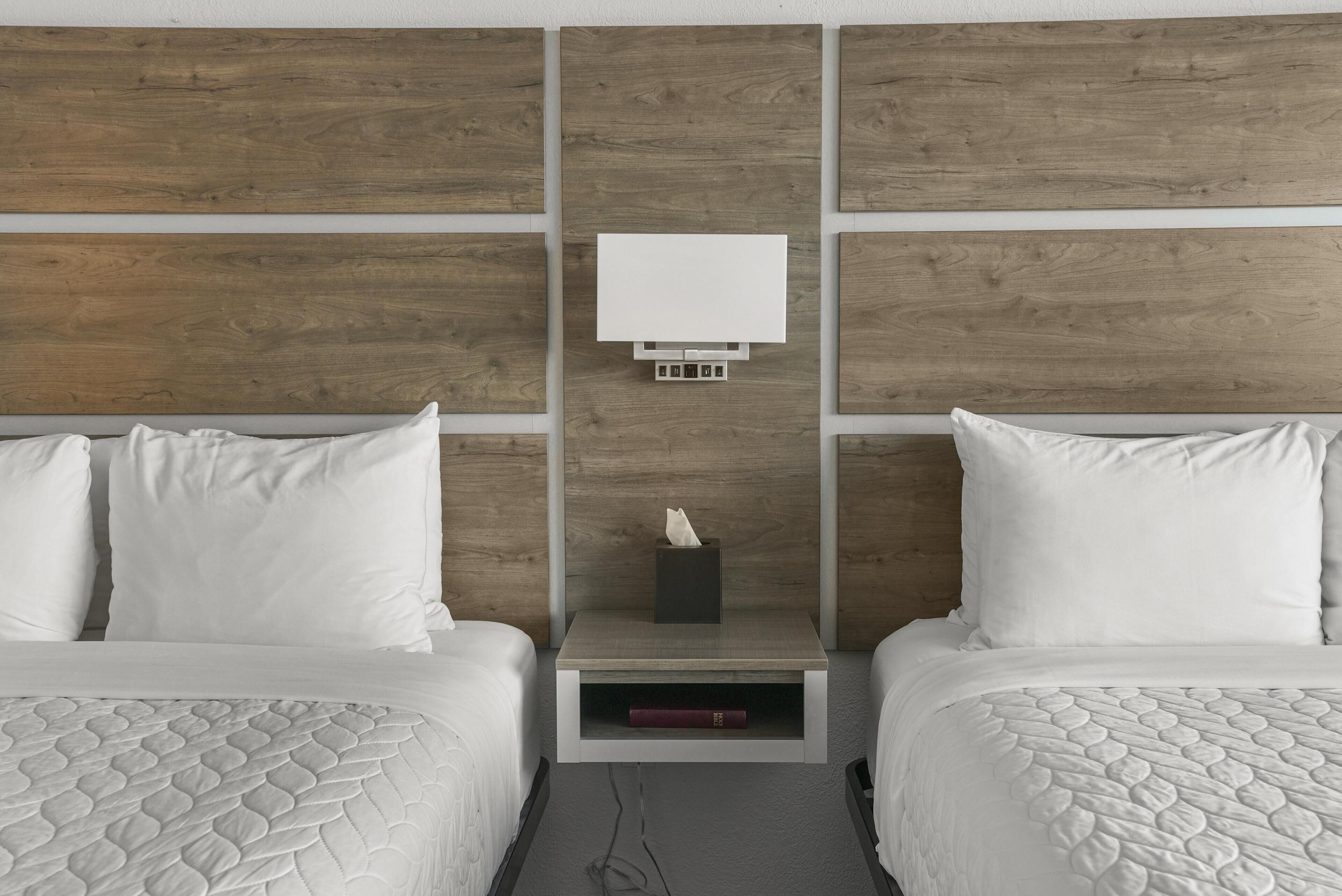 Habitación de hotel con dos camas, una mesita de noche con lámpara y cabecero de madera.