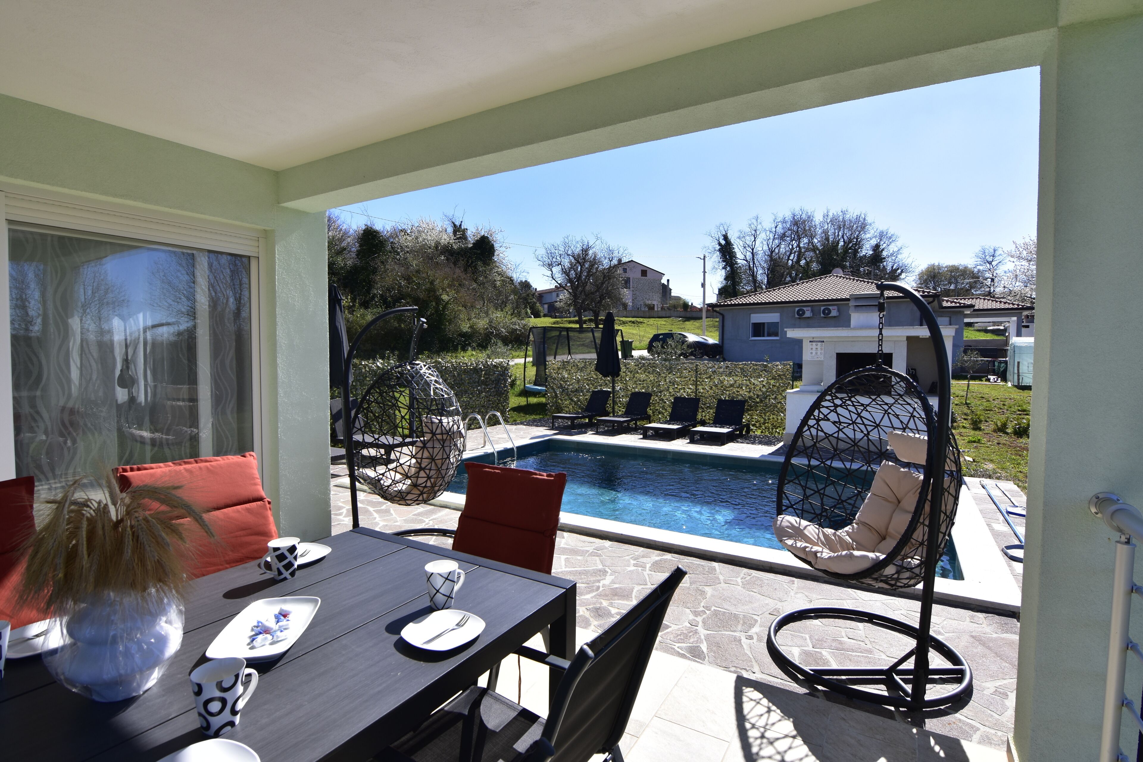 Une terrasse avec une table à manger, des chaises et un fauteuil suspendu, le tout à proximité d’une piscine.