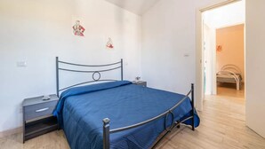 Une chambre à coucher avec un lit en métal, une couvre-lit bleue, une table de chevet et une commode. Une porte donne accès à une autre pièce.